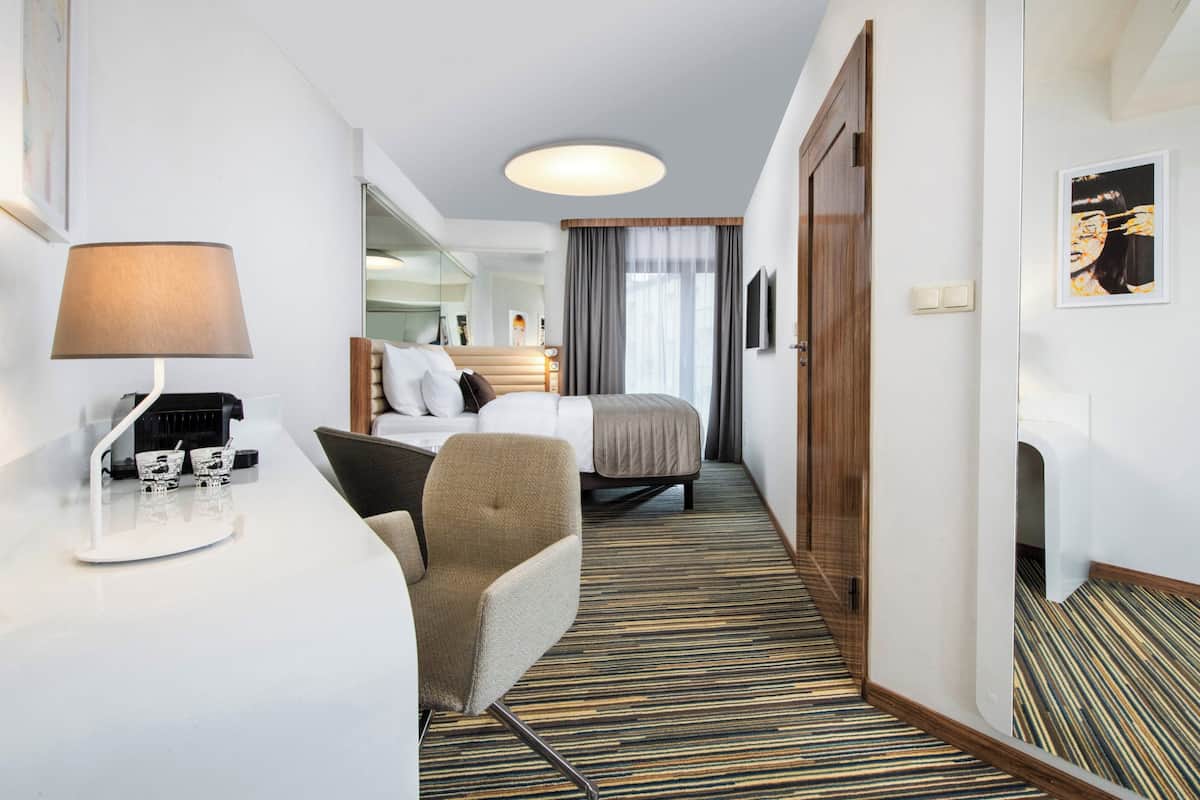 Hotellihuone, jossa on sänky, kirjoituspöytä lampun kanssa, tuoli ja peili.