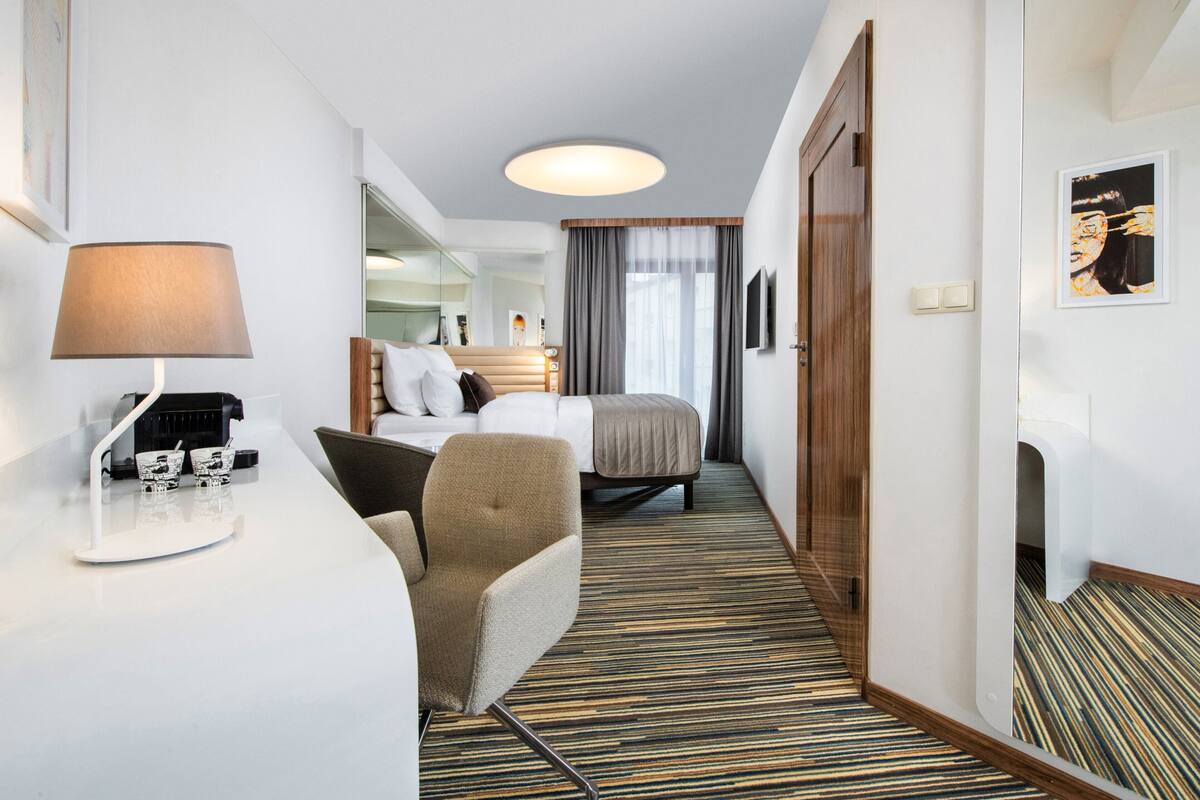 A hotel room with a bed, a desk with a lamp, a chair, and a mirror.