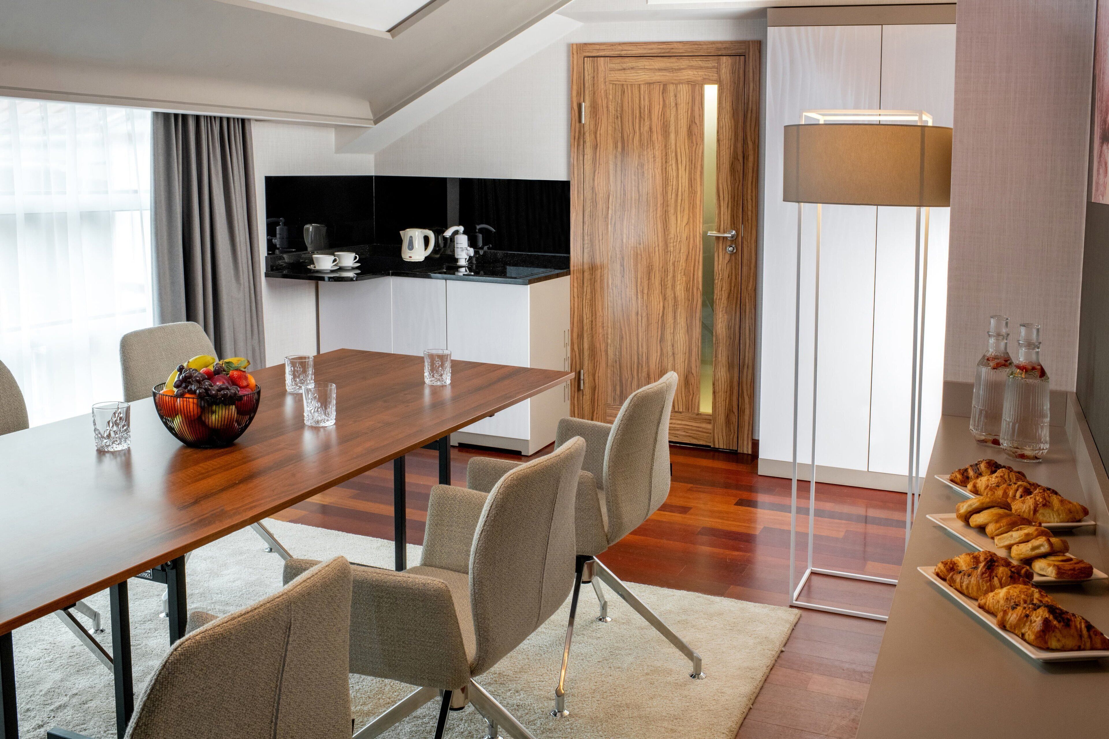 Una habitación de hotel moderna con una mesa de comedor de madera, sillas cómodas, un frutero, una zona de cocina con cafetera y un ventanal grande.