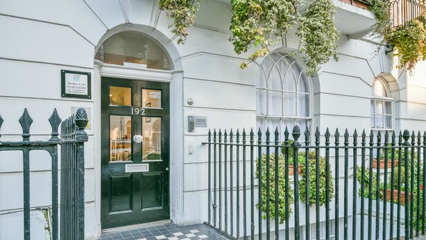 A building entrance with a black and white checkered floor, a black door with the number 192, and a black iron fence with greenery.