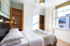 Una habitación de hotel con una cama, una estantería de madera, una ventana con cortinas y una lámpara en el techo.