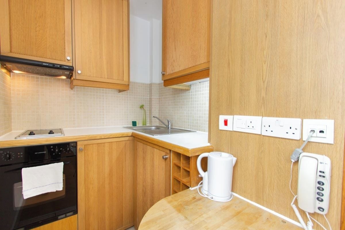 Una cocina con armarios de madera, un horno negro, un lavamanos blanco y una tetera blanca sobre la encimera.