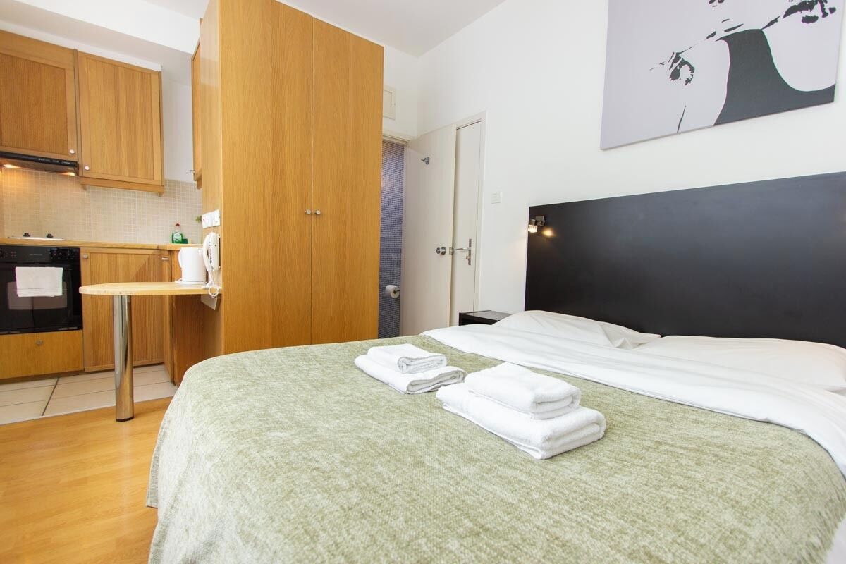 A hotel room with a bed, a small table, a kitchen area, and a door.