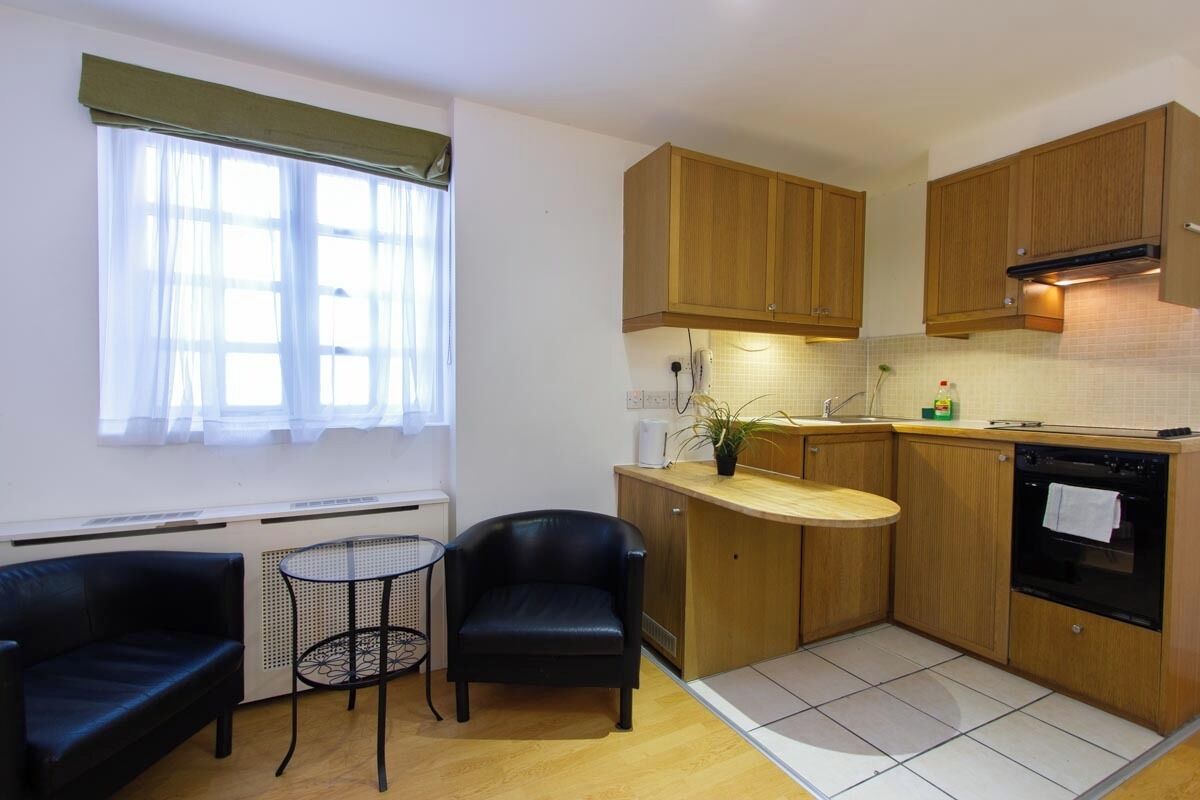 Cocina compacta con armarios de madera, una pequeña mesa de comedor, dos sillas y un ventanal con cortinas de gasa.
