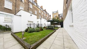 A narrow alleyway with a small garden area, a white building, and a bench.