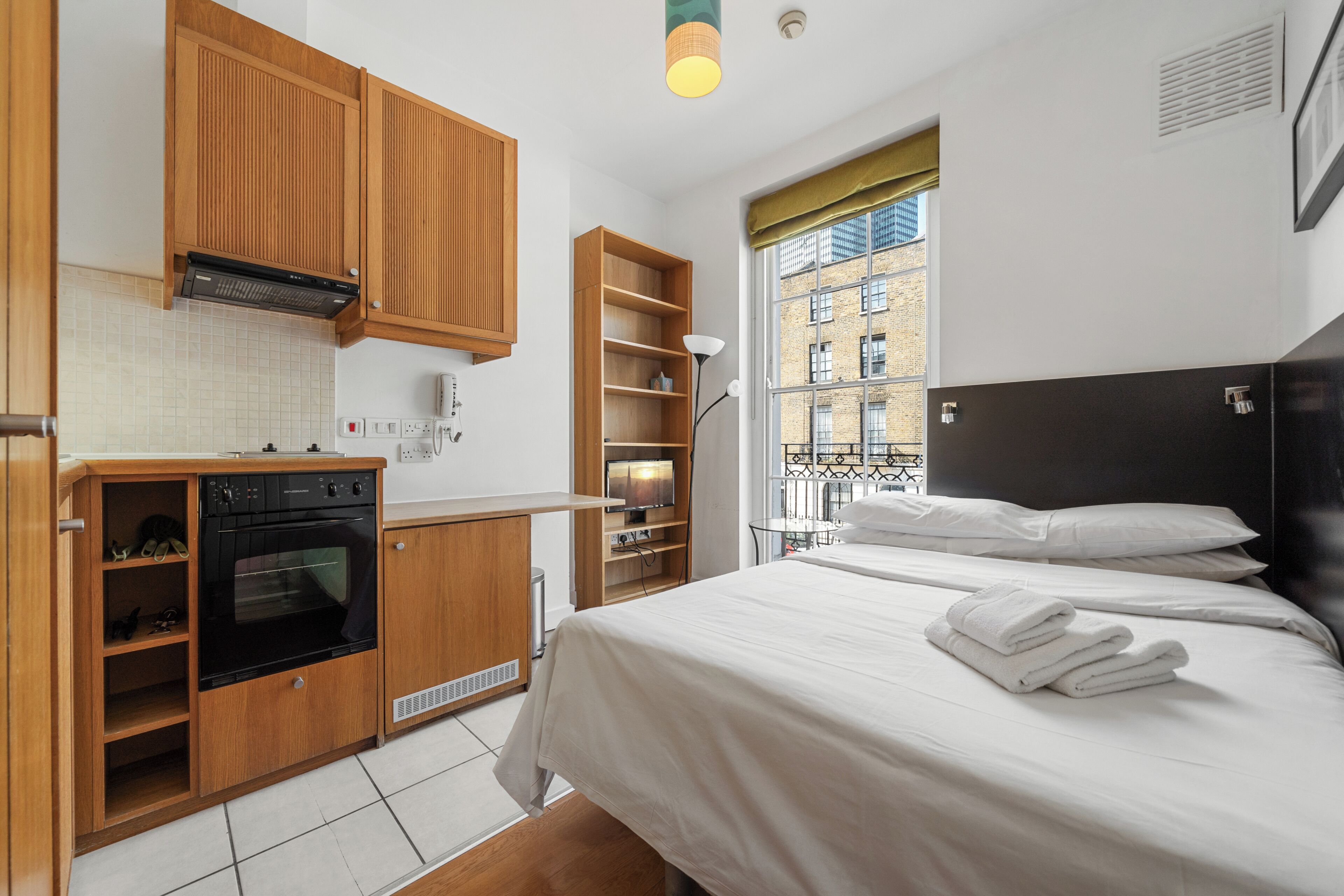 A compact bedroom with a bed, a small kitchenette, and a window with a view of buildings.