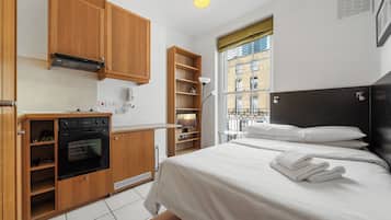 Un dormitorio compacto con una cama, una kitchenette pequeña y una ventana con vistas a edificios.