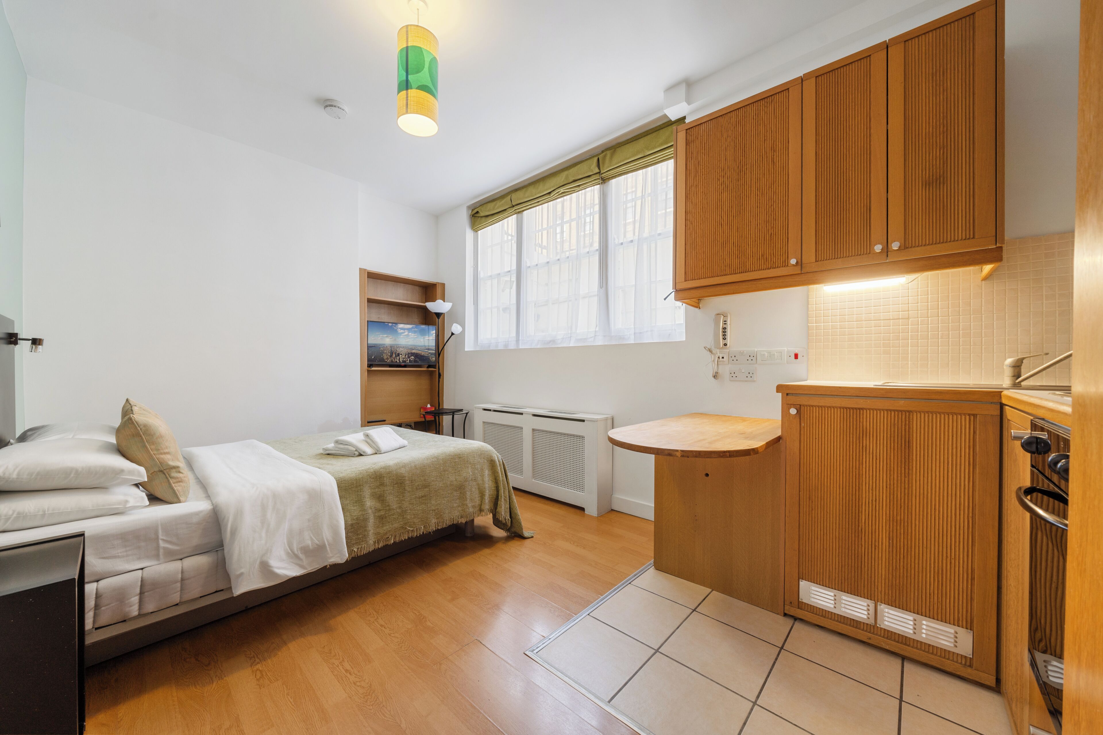 A compact room with a bed, a small table, a window with curtains, a radiator, and wooden cabinets.