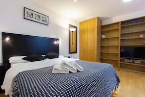 Habitación de hotel con una cama grande, un armario, televisor y estante con cestas.