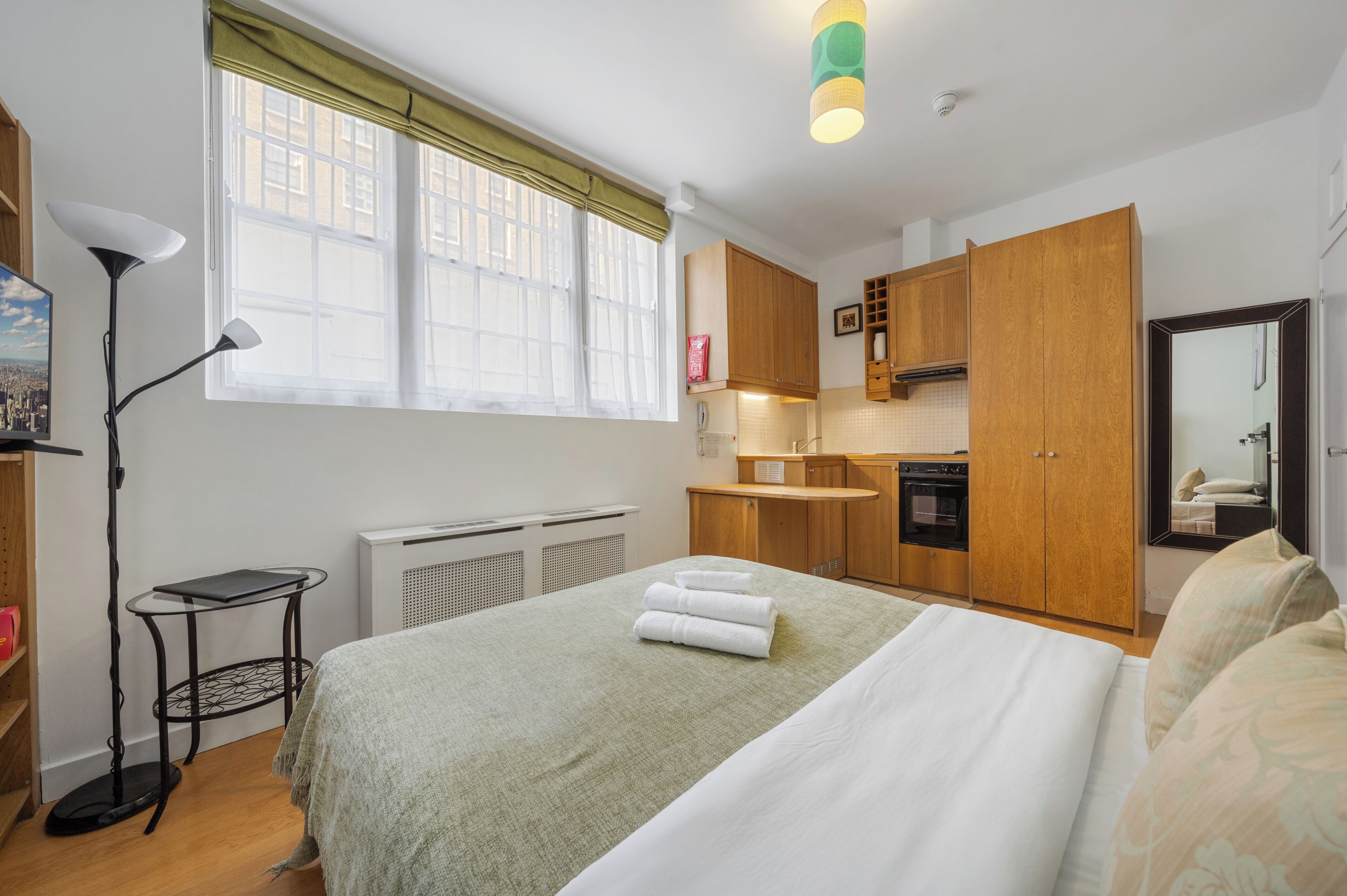 Une chambre d’hôtel comprenant un lit, un bureau, une chaise, un radiateur, une fenêtre avec des rideaux, un coin cuisine avec des placards et une cuisinière, et un miroir.