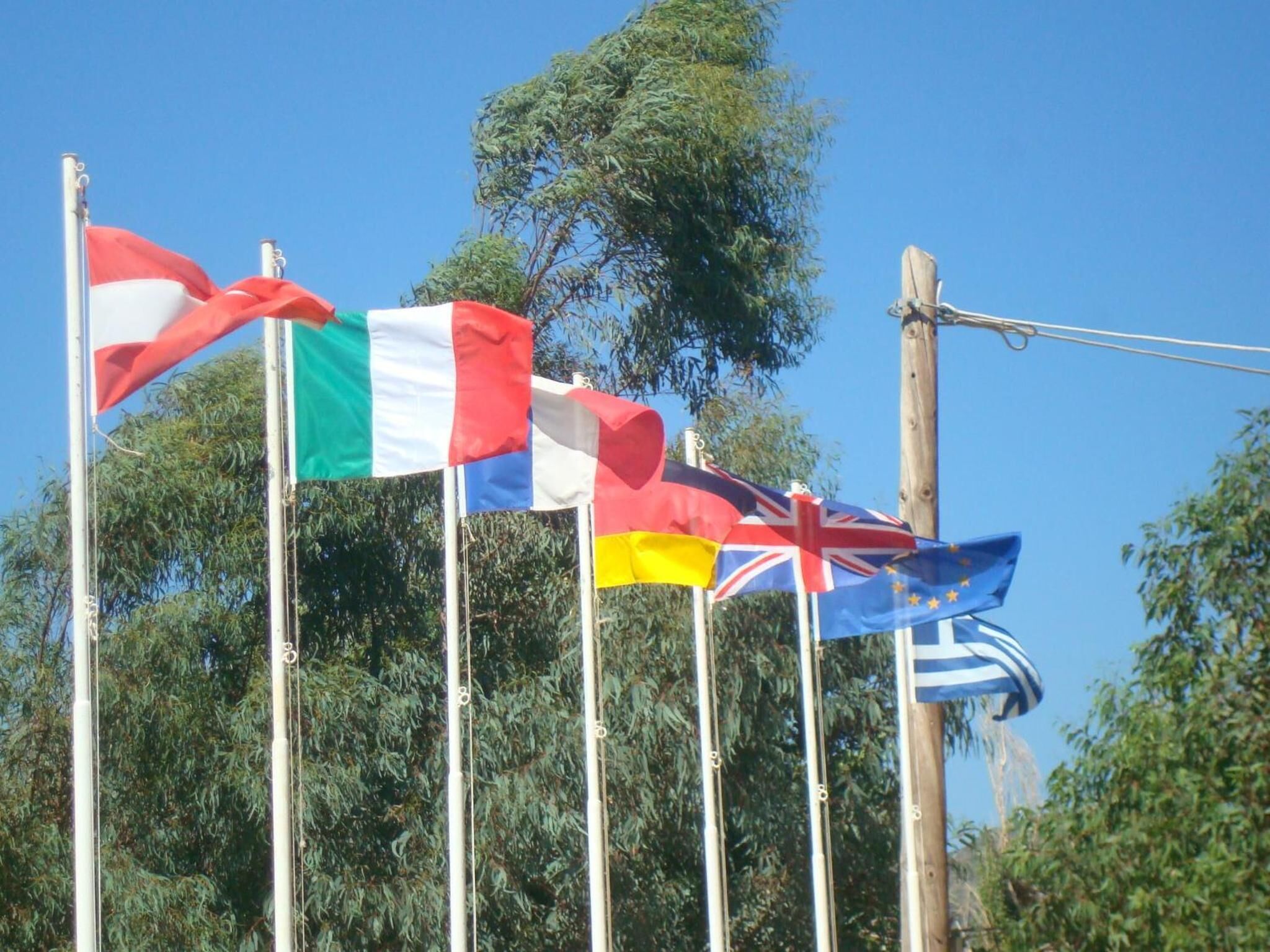 Een rij vlaggen op vlaggenmasten, waaronder de Italiaanse vlag, de Britse vlag en de Europese vlag.