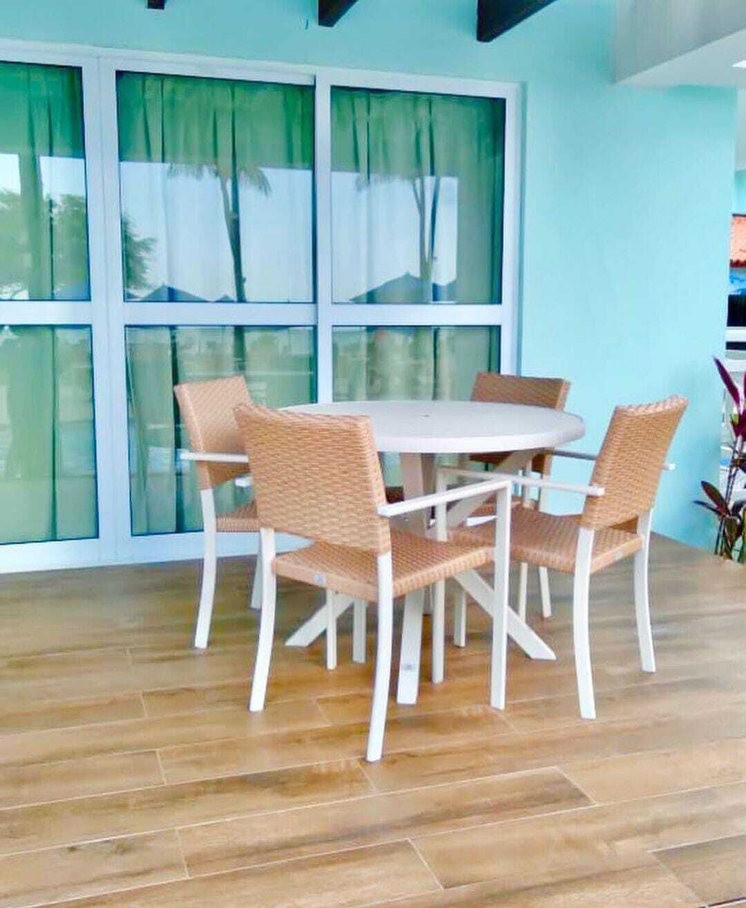 A dining area with a round table and six wicker chairs, large windows with curtains, and a wooden floor.
