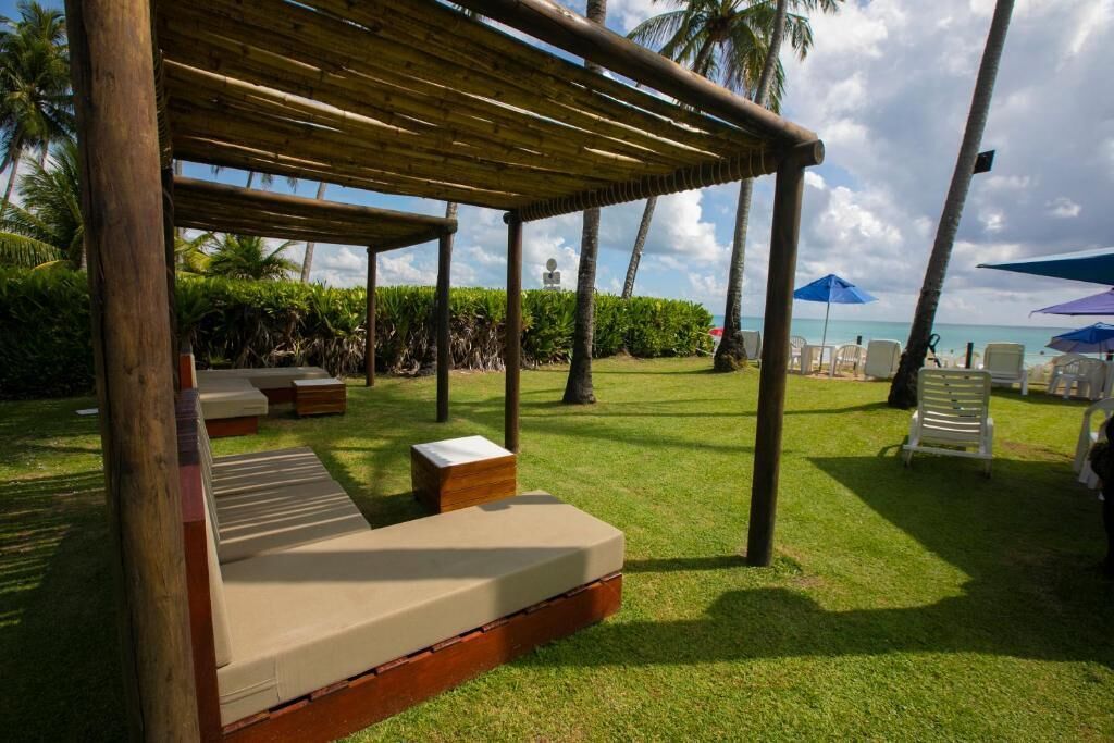 Un'area relax all'aperto con tettoia in legno, panche imbottite e vista sulla spiaggia e sulle palme.