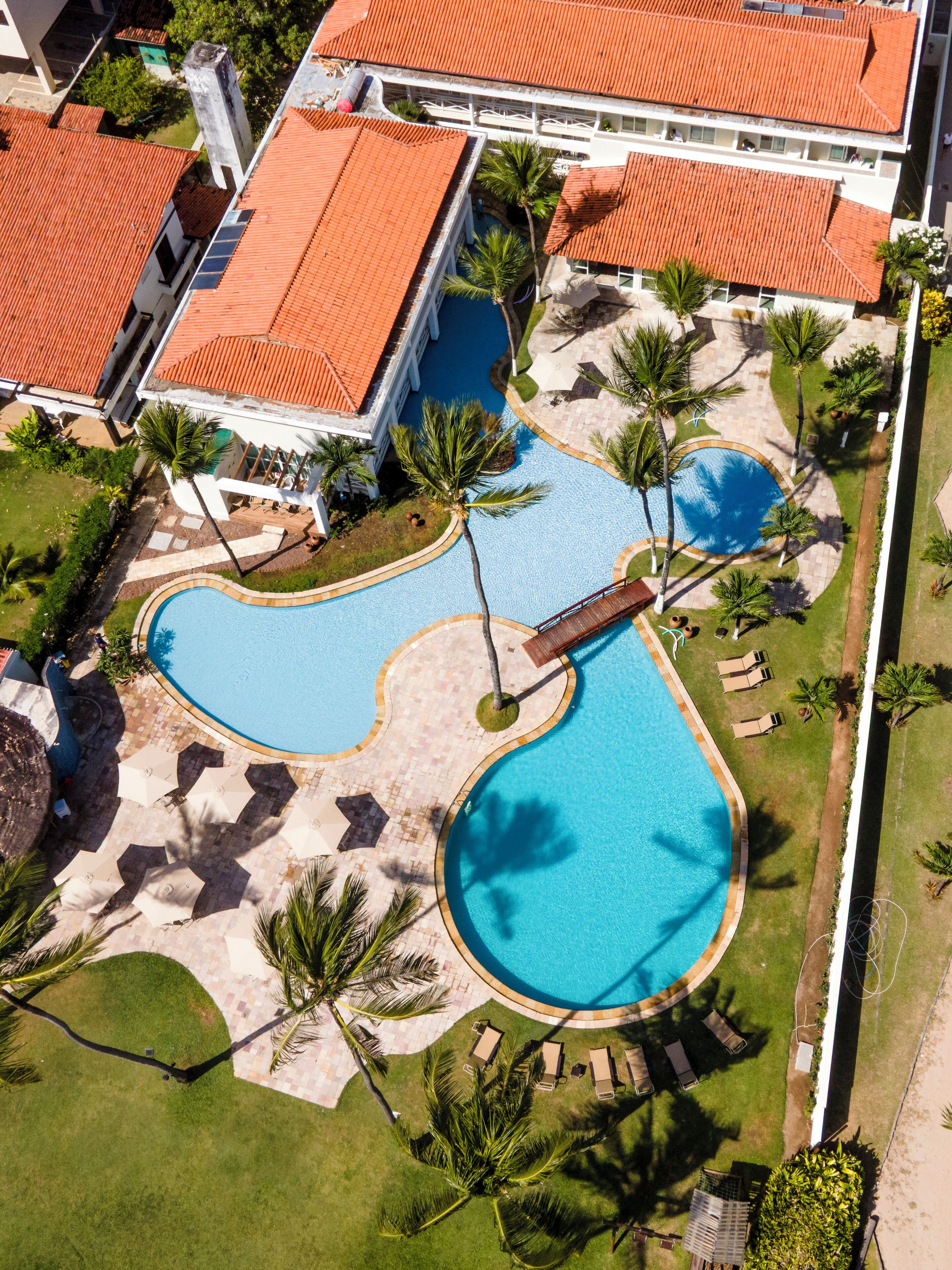 Vista aérea de un resort con dos piscinas, palmeras y un edificio de techo rojo.