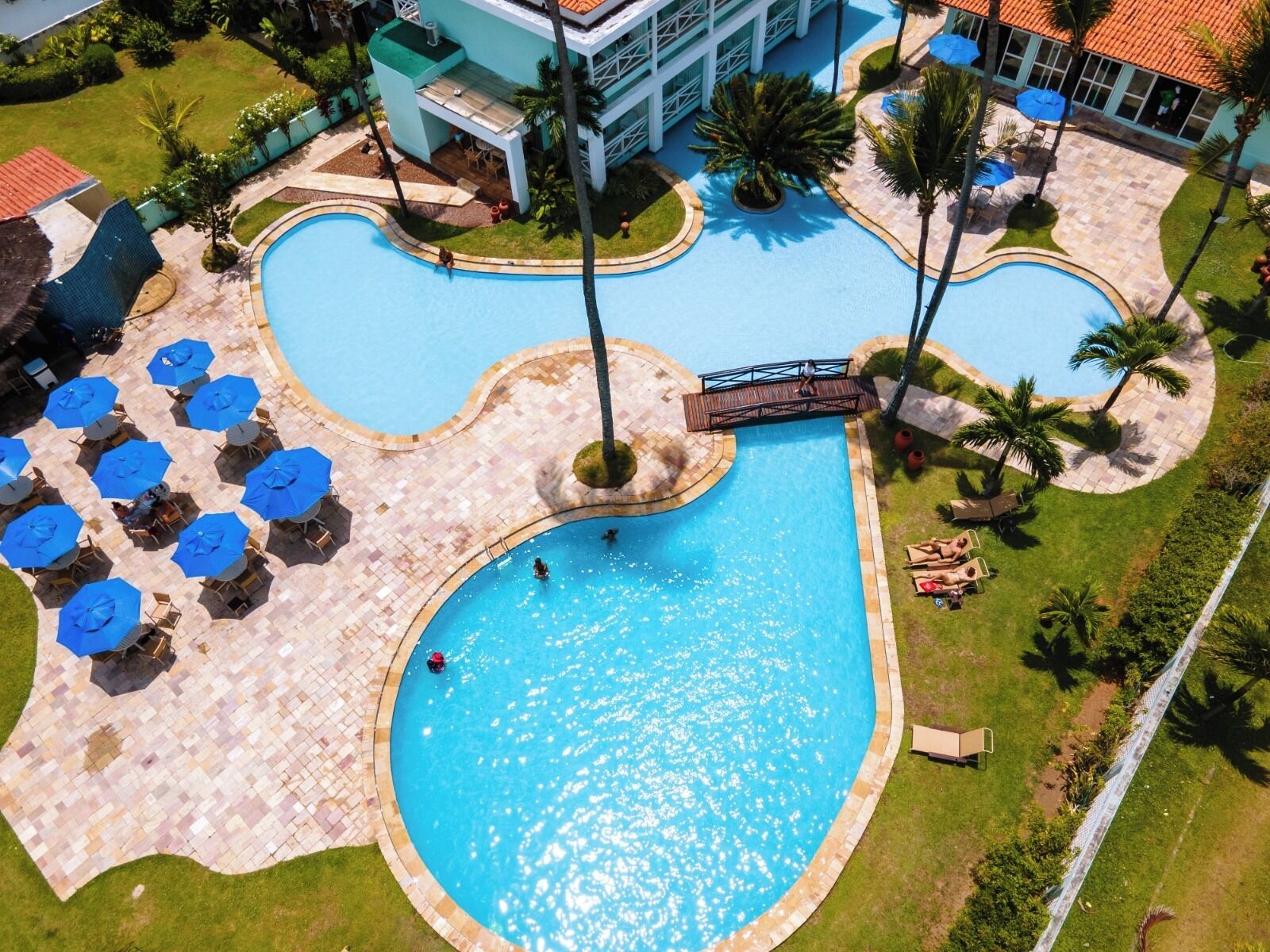 An aerial view of a resort with a central swimming pool, lounge areas with umbrellas, and palm trees.