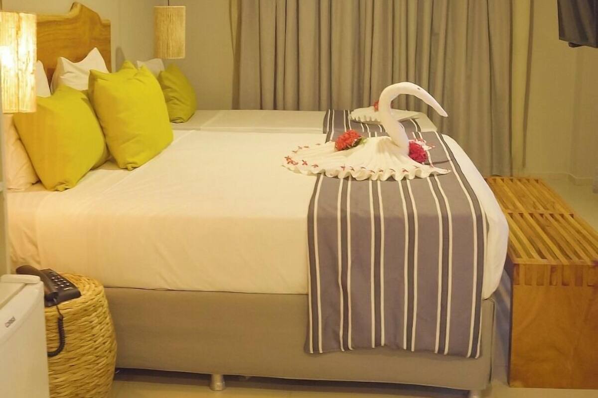 Habitación de hotel con cama, almohadas, mesita de noche, lámpara, zapatero y cortina.