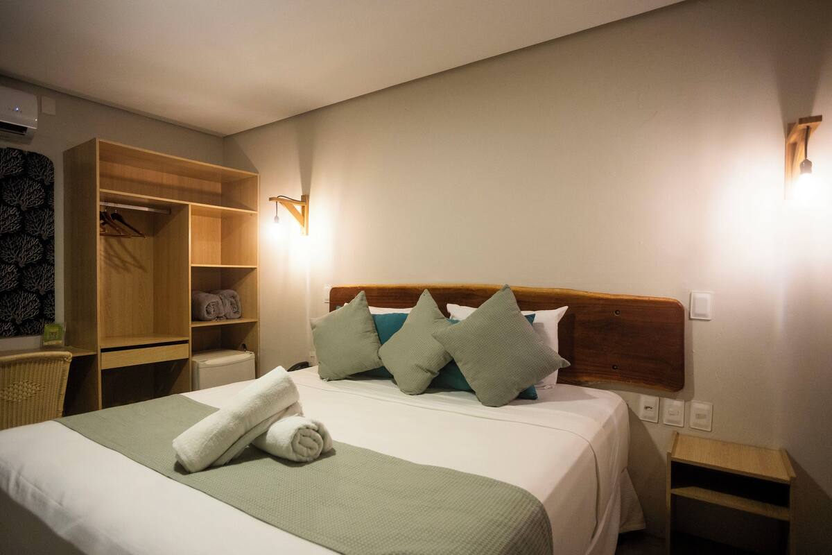 Habitación de hotel con una cama, un armario de madera, una mesita de noche y un aplique de pared.