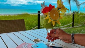 A hand holding a cocktail with fruit garnish, a book, and a smartphone on a table with a view of the ocean and palm trees. - Baia Branca Beach Resort (Tamandaré)