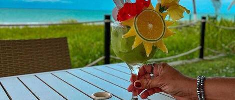 A hand holding a cocktail with fruit garnish, a book, and a smartphone on a table with a view of the ocean and palm trees.