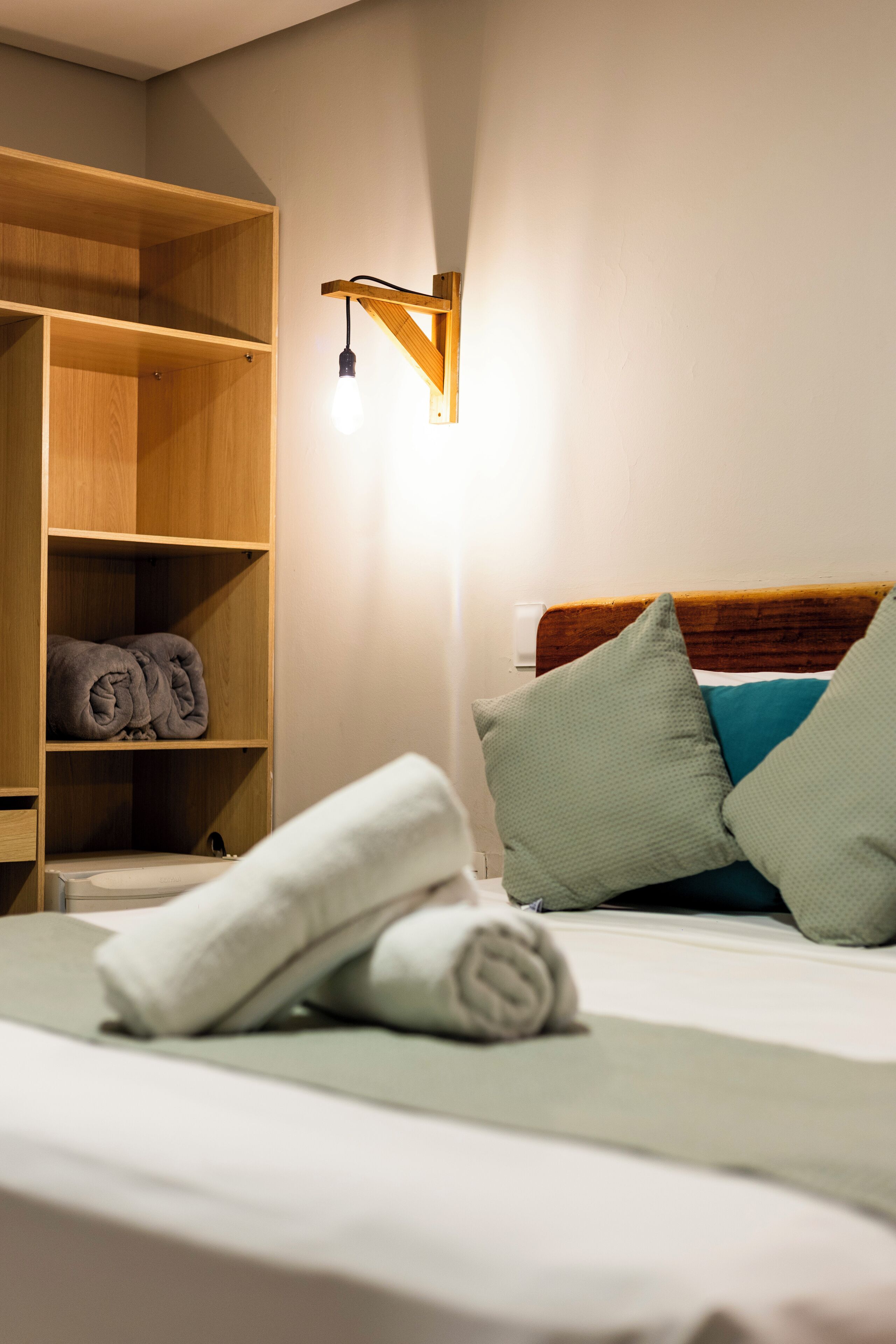 A neatly made bed with pillows and a rolled towel, a wooden wardrobe, and a wall-mounted light fixture.