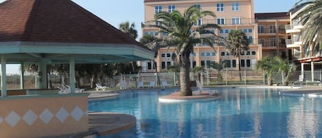 A swimming pool with lounge chairs and a gazebo, surrounded by palm trees and a multi-story building with balconies.