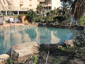Una piscina con borde de piedra, rodeada de vegetación y un edificio con balcones.