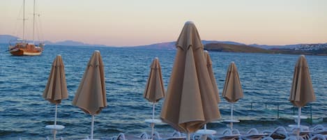 A beach with lounge chairs and umbrellas, a sailboat in the background, and a calm sea.