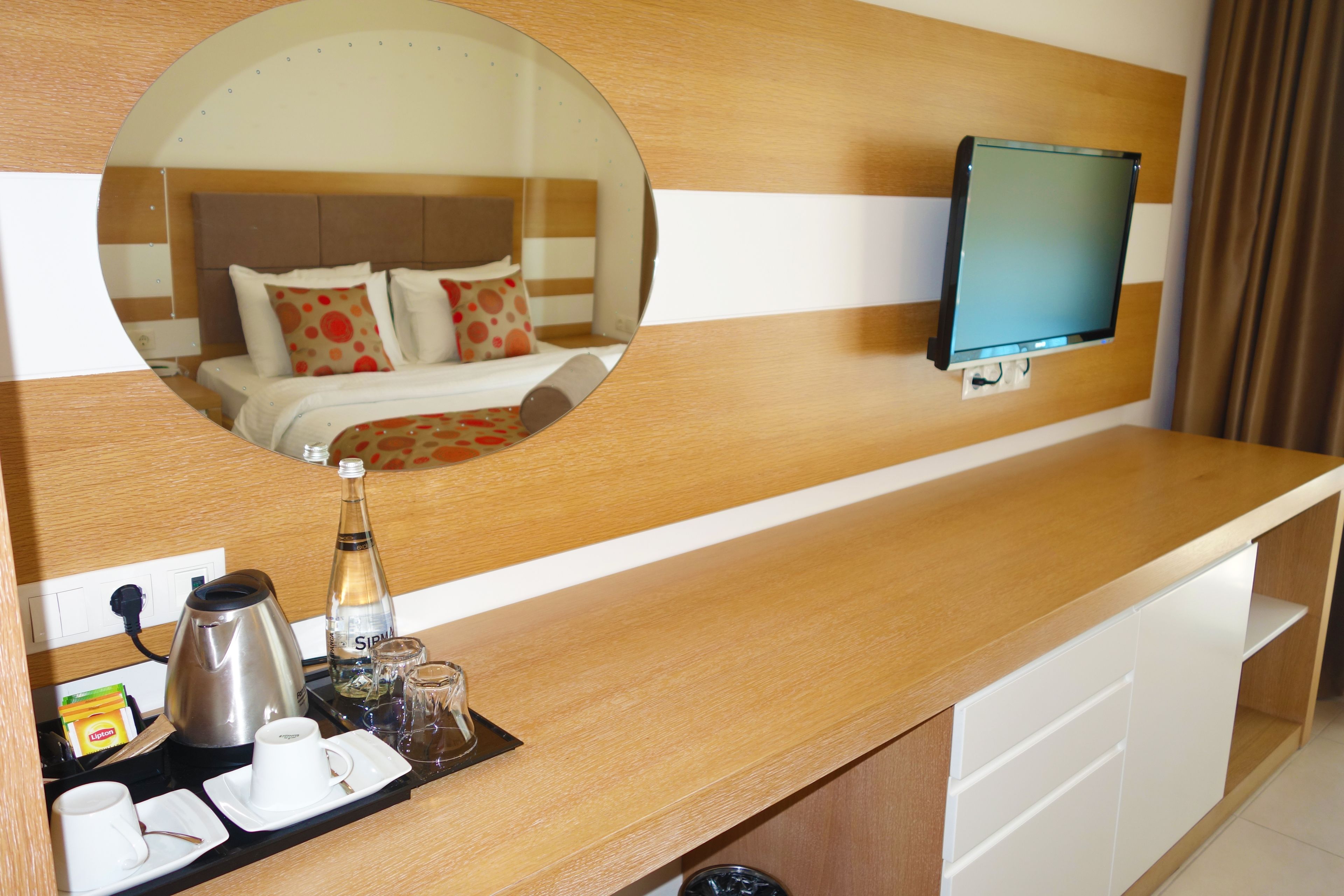 Habitación de hotel con mostrador de madera, un espejo que refleja la cama, un televisor montado en la pared y una cafetera sobre el mostrador.