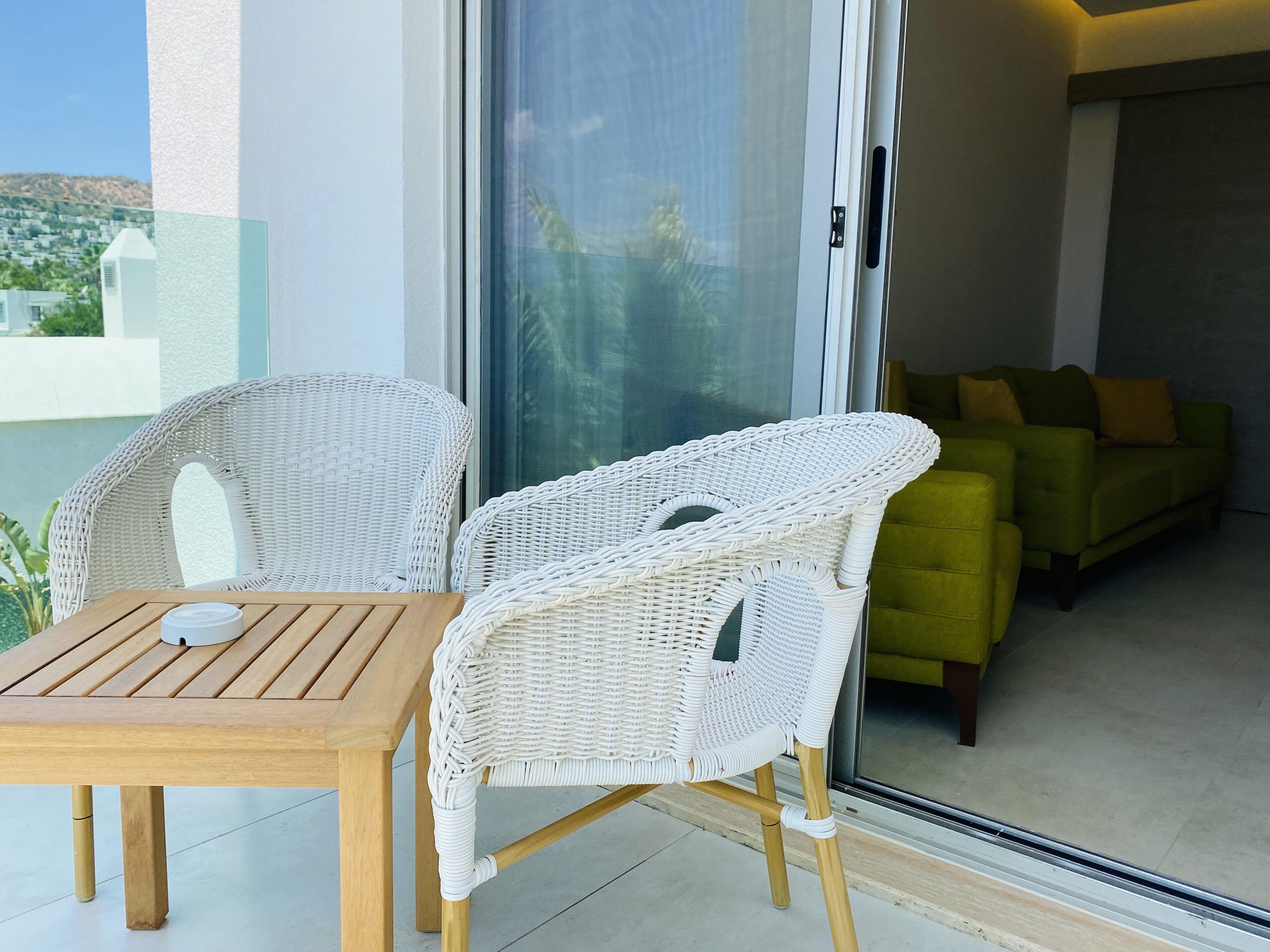 Un balcón con mobiliario de mimbre, una mesa de madera y un ventanal corredero que da a un interior con un sofá verde.