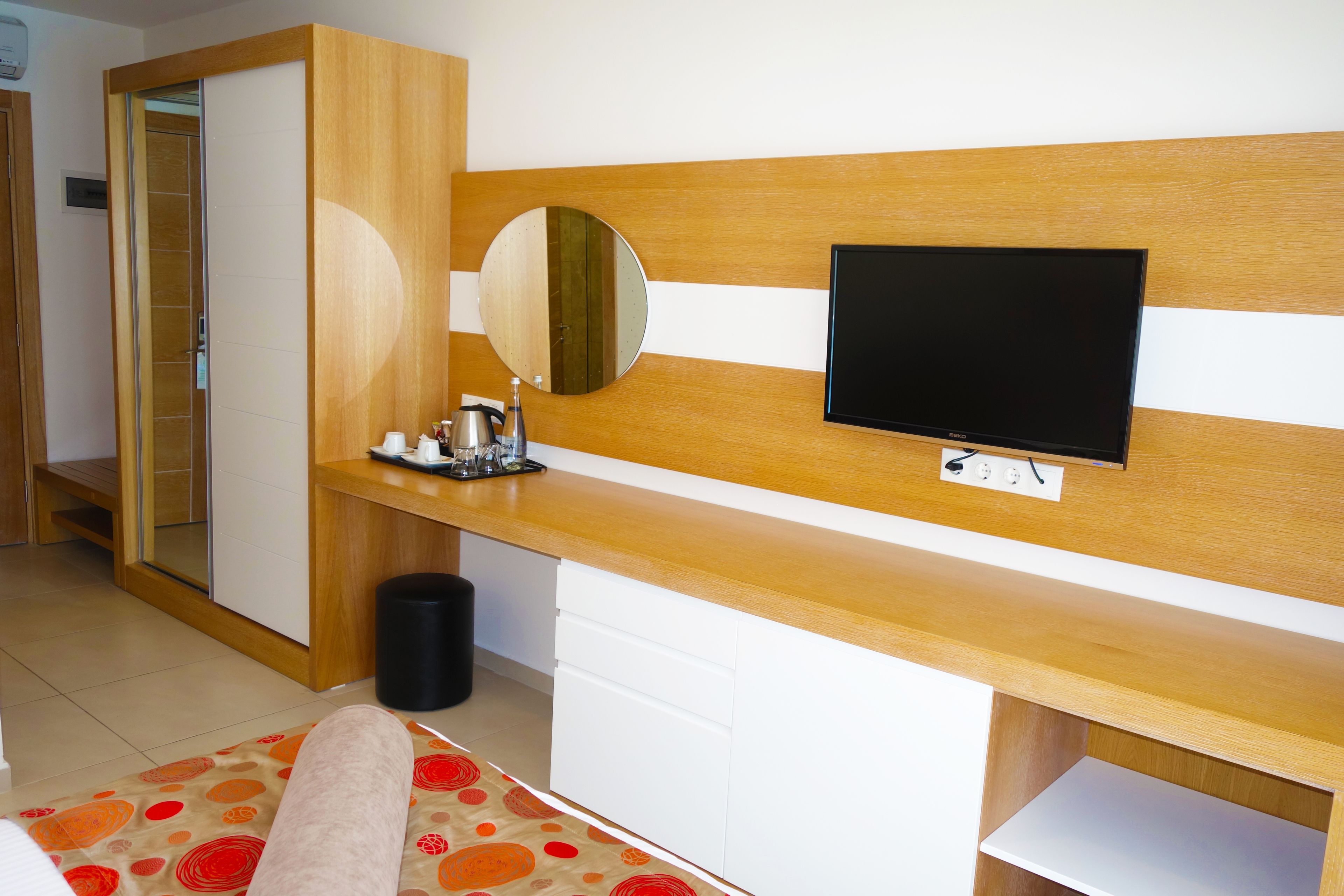 A hotel room with a wooden headboard, a flat-screen TV mounted on it, a bedside table with a mirror, a small trash bin, and a bed with a patterned bedspread.