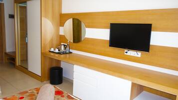 A hotel room with a wooden headboard, a flat-screen TV mounted on it, a bedside table with a mirror, a small trash bin, and a bed with a patterned bedspread.