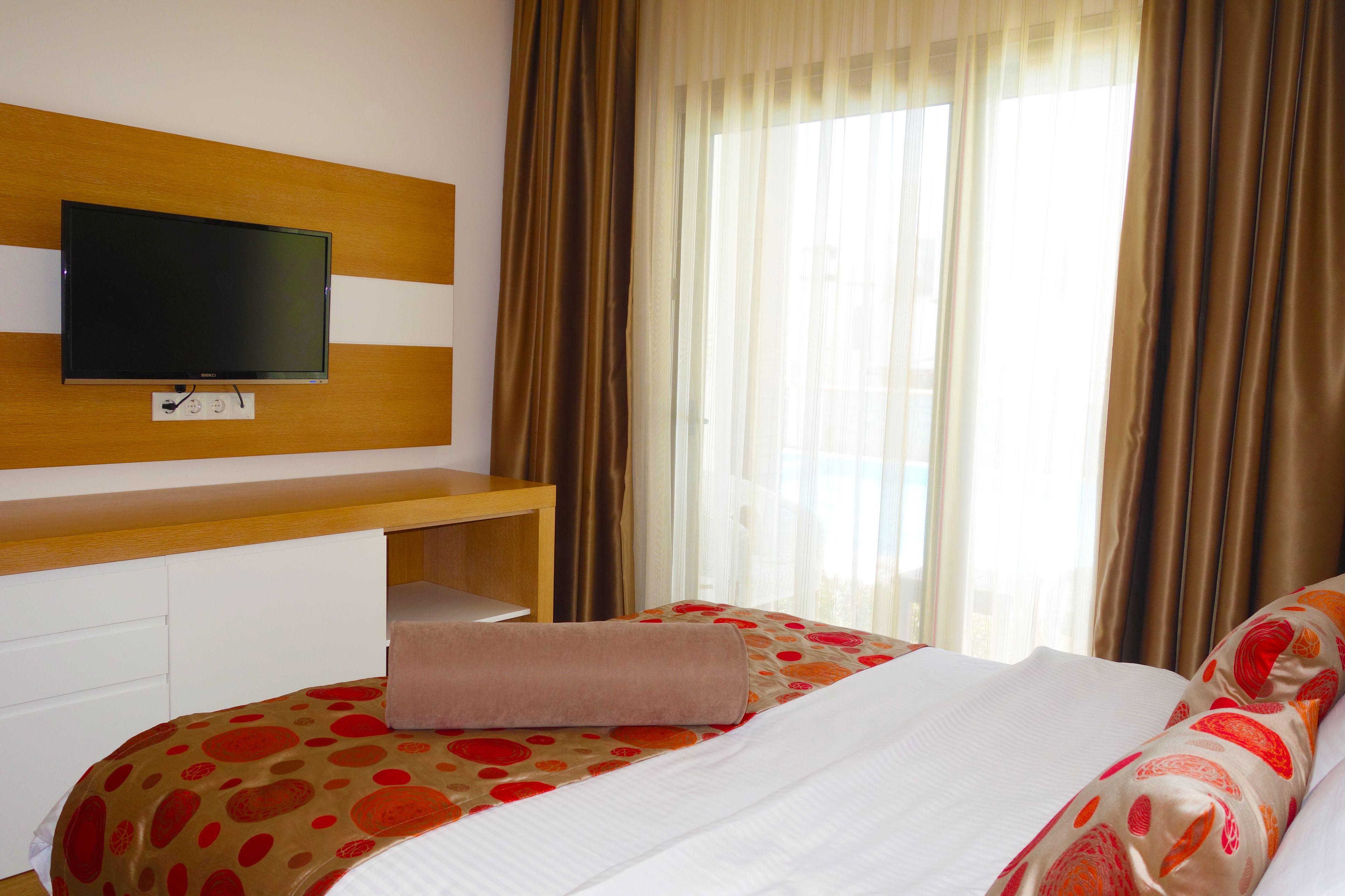 A hotel room with a bed, a TV mounted on the wall, a desk, and a window with curtains.