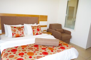 Una habitación de hotel con una cama grande, un cabecero marrón, un sillón marrón y una ventana con persianas.