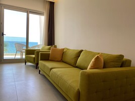 Une pièce avec un canapé vert, une chaise blanche et une vue sur la mer, visible à travers une porte coulissante.