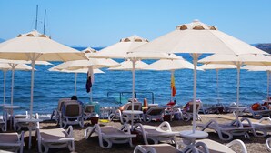 Un cadre idyllique en bord de mer, avec de nombreux fauteuils de plage blancs et des parasols beiges, disposés sur une zone sablonne offrant une vue sur la mer.