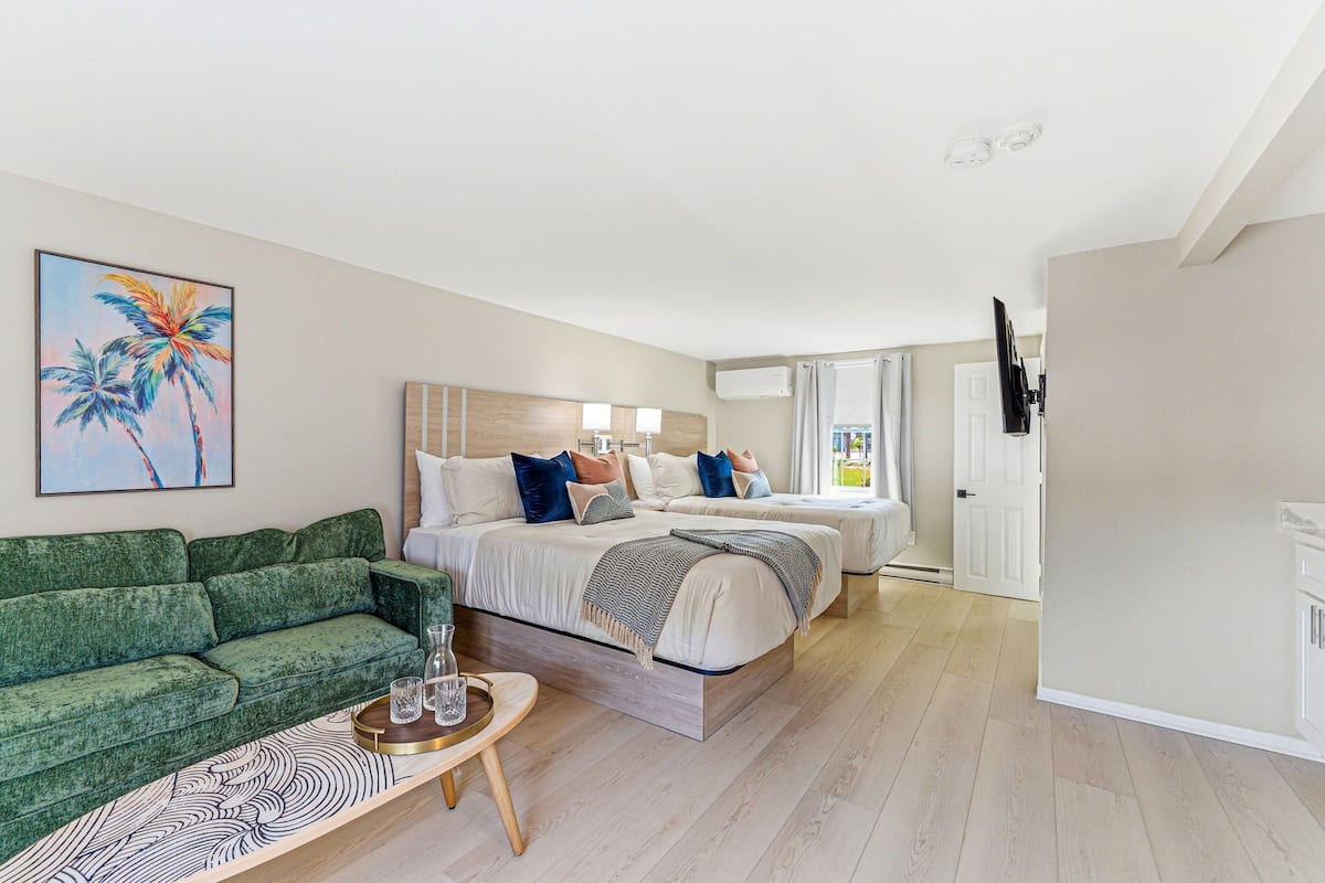 Habitación de hotel con cama, sofá verde, una mesita con vasos y un cuadro de palmeras.