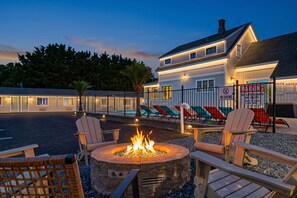 A fire pit surrounded by Adirondack chairs, a pool area with lounge chairs, and a building with a 'No Diving' sign.