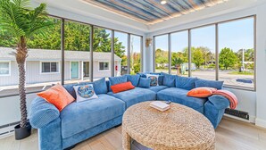 A modern living room with a large blue sectional sofa, a woven coffee table, and a large window offering a view of the street. - Dennis Port Surf Club (Boutique Hotel) (Dennis Port)