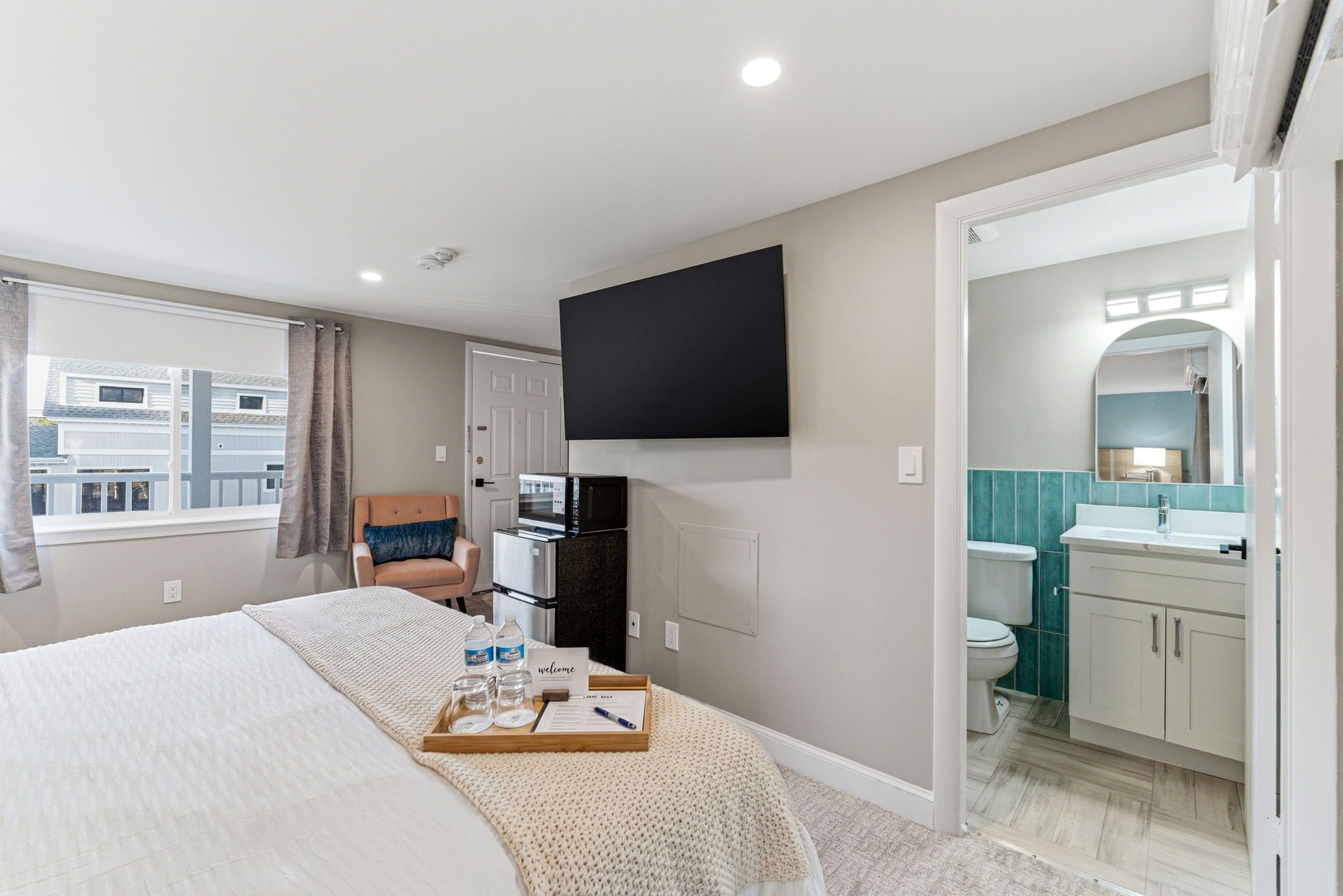 A bedroom with a bed, a TV mounted on the wall, a chair, a small kitchenette, and a bathroom visible through an open door.