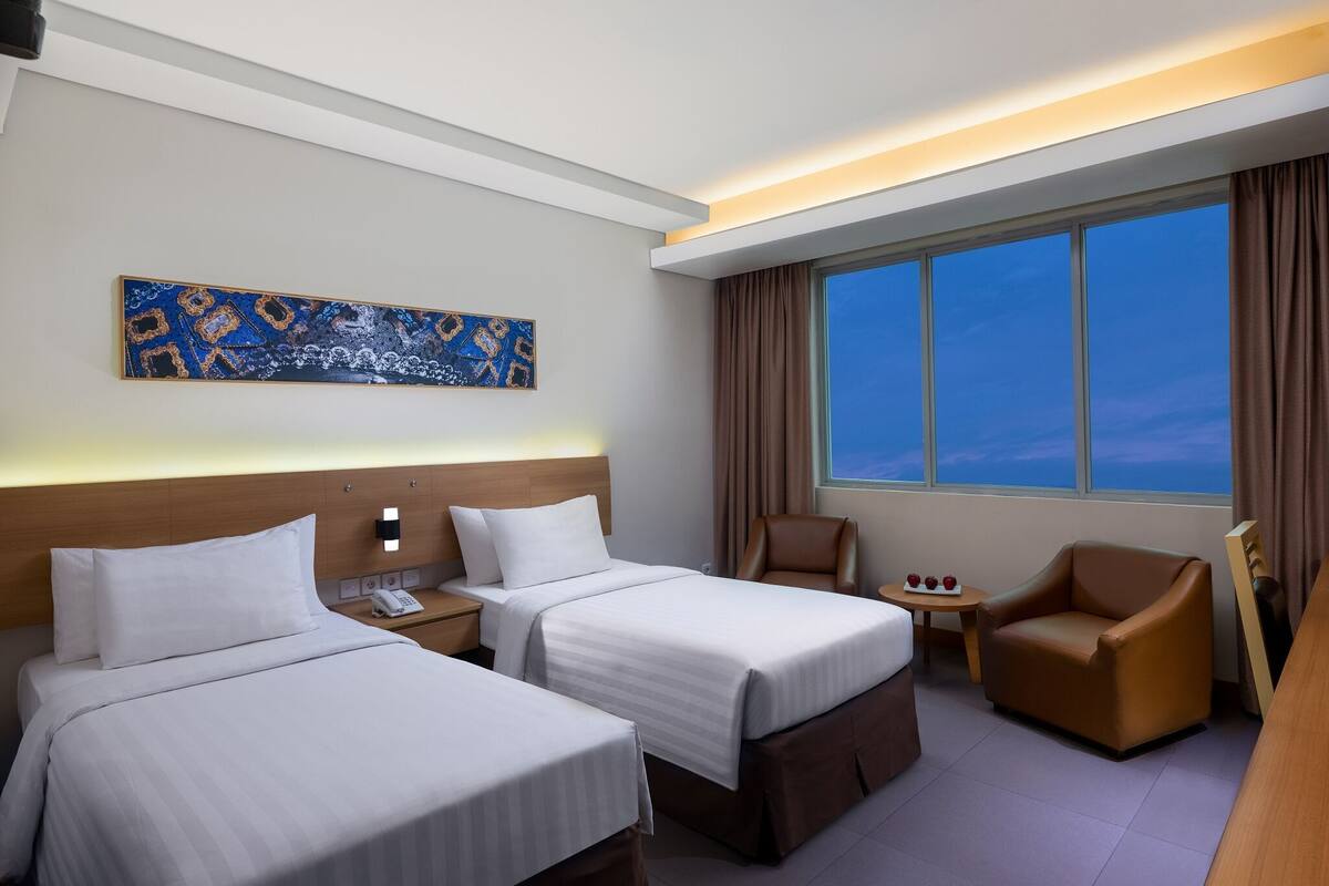 A hotel room with two beds, a desk, a chair, a painting on the wall, and a large window.