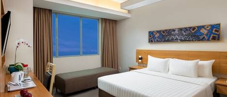 A hotel room with a large bed, a desk with a chair, a small sofa, a nightstand, a lamp, and a window with curtains.