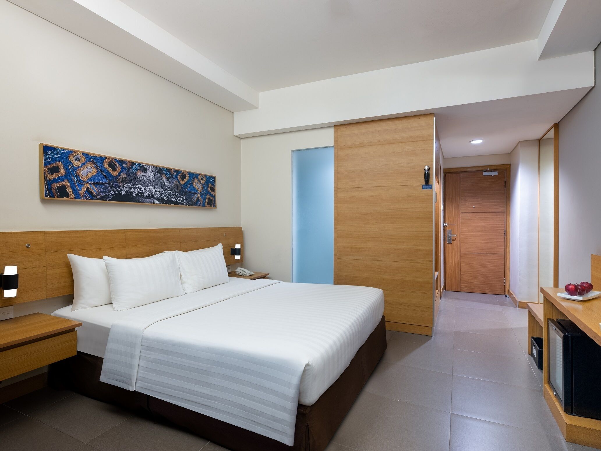 A modern hotel room with a large bed, a nightstand, a desk with a computer, and a door leading to another room.