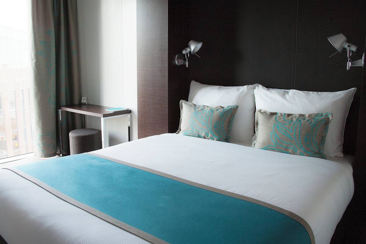 Een hotelkamer met een groot bed, twee bedlampen, een klein tafeltje en uitzicht op de stad door het raam.