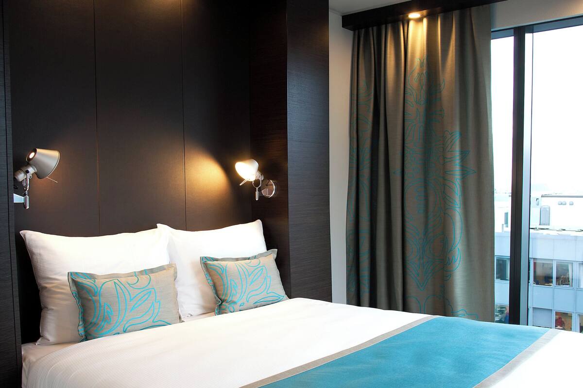 Hotelkamer met een groot bed, twee wandlampen en uitzicht op de stad door het raam.