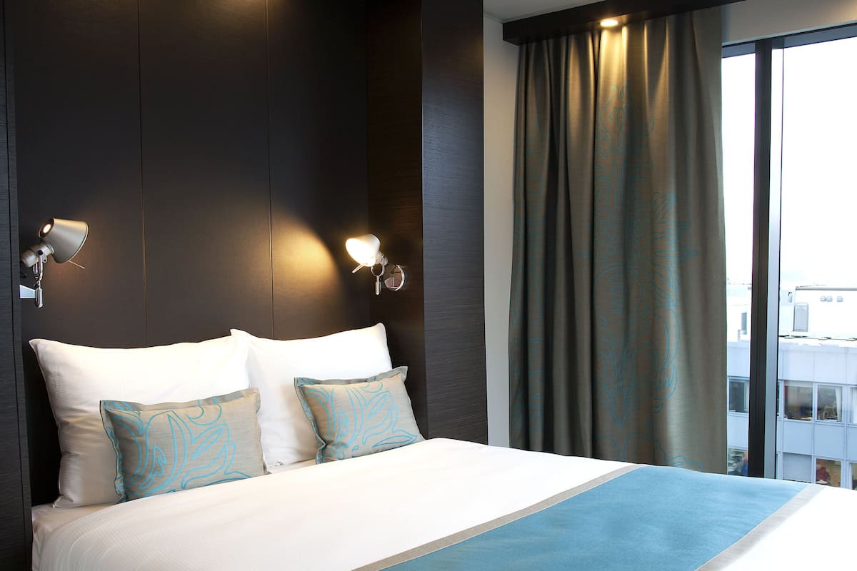 Ein Hotelzimmer mit einem großen Bett, zwei Wandlampen und Blick auf die Stadt durch das Fenster.