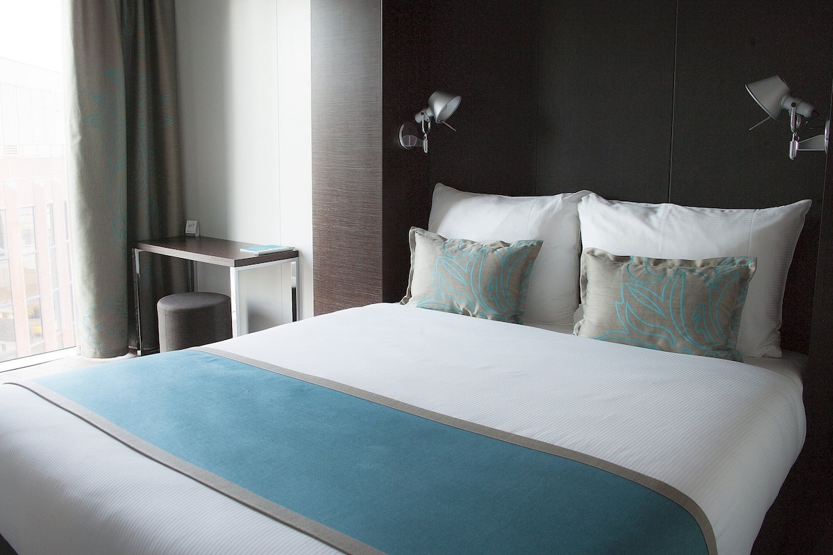 Ein Hotelzimmer mit einem großen Bett, zwei Nachttischlampen, einem kleinen Tisch und Blick auf die Stadt durch das Fenster.