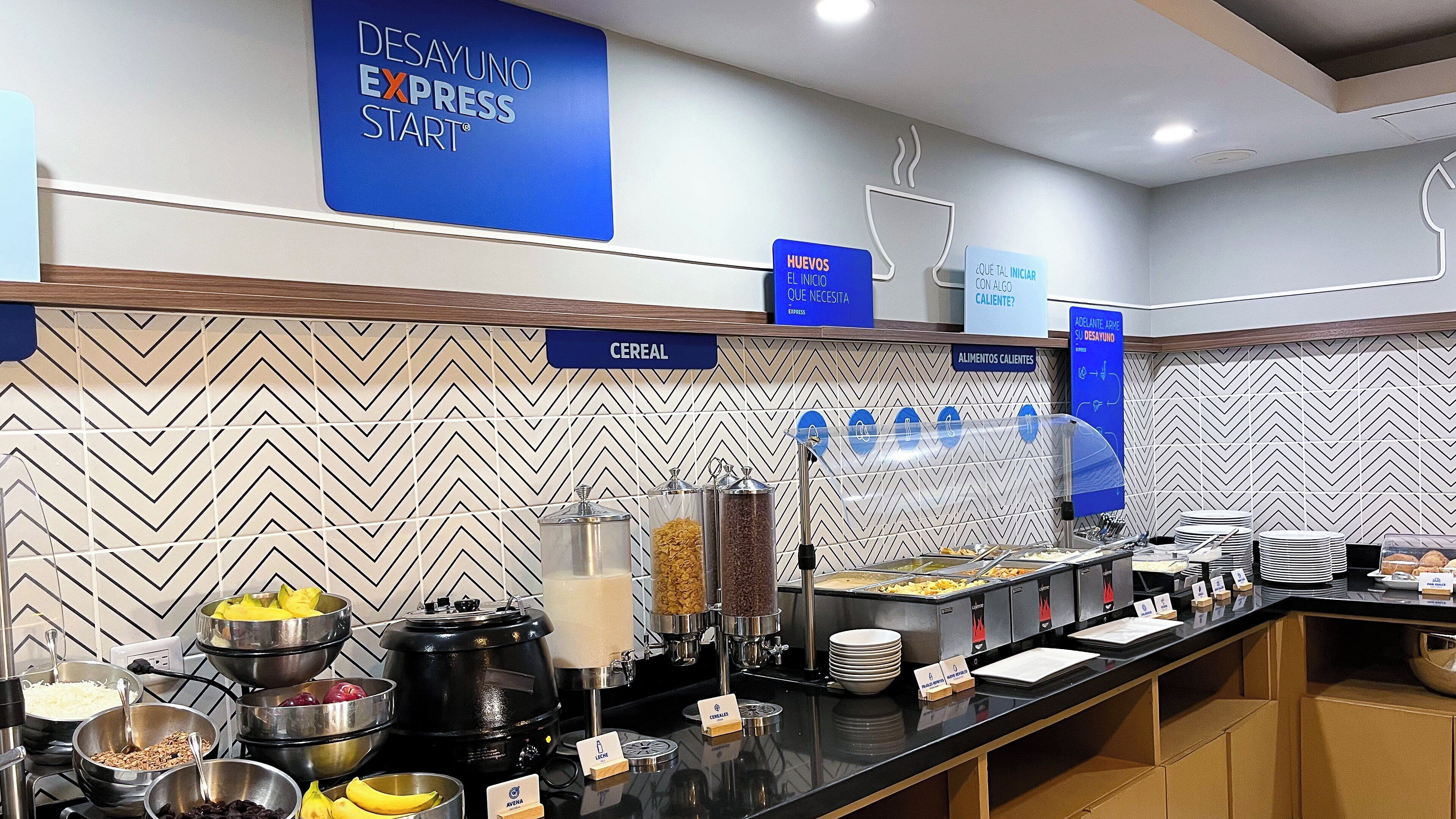 Zona de desayuno tipo buffet con secciones identificadas para cereales, alimentos calientes y bebidas.