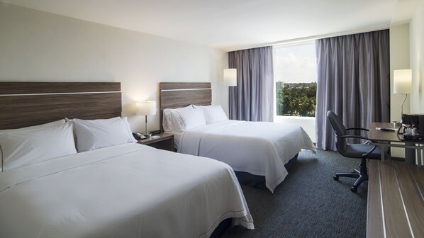 Ein Hotelzimmer mit zwei Betten, einem Schreibtisch mit Stuhl, einer Lampe und Blick auf Palmen durch das Fenster.