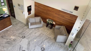 Un área de recepción con mostrador de madera, dos sofás grises, una mesa con un arreglo floral y un suelo de mármol con señalización.