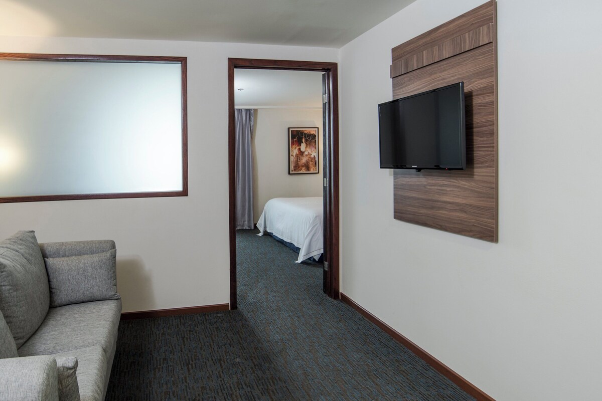 A hotel room with a sofa, a flat-screen TV, a large mirror, and a door leading to a bedroom with a bed and a painting.